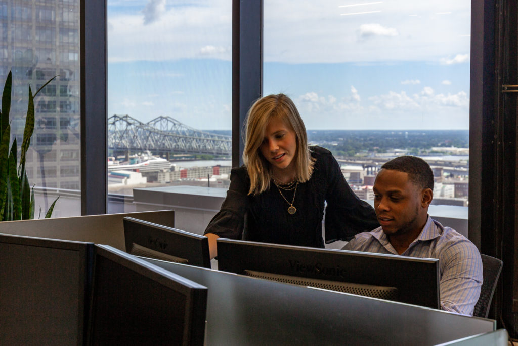 A man and woman sit at a computer together with large windows overlooking downtown New Orleans